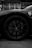 A black and white photograph focusing on the front wheel of a sports car. The car has a sleek body with a prominent badge on the side panel. The tire and hubcap are detailed, showcasing a stylish design with visible brake components. A person is faintly visible in the background standing near a wall.