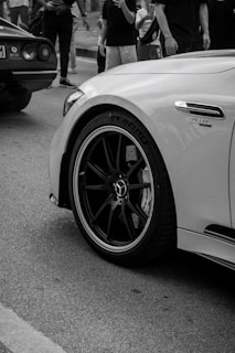 A high-performance car is parked on the street surrounded by people. The focus is on the front wheel, bearing the logo of a well-known luxury car brand. A badge reading 'Turbo' is visible on the side of the car. Another car is partially visible in the background. The setting appears to be a public gathering or event, as several individuals are standing and capturing photos.