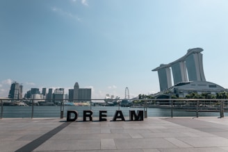 The scene depicts a waterfront cityscape with a prominent modern building featuring three towers connected by a sky park. In the foreground, large letters spell out the word 'DREAM' on a paved area. Several other skyscrapers are visible across the water, and a Ferris wheel stands out in the background. The sky is clear with a few clouds.