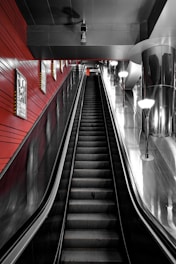 An indoor escalator ascends in a modern space, with sleek metallic surfaces and red walls. Bright overhead lights illuminate the area, reflected in the shiny metal surfaces. Posters are mounted on the walls flanking the escalator.