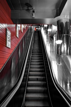An indoor escalator ascends in a modern space, with sleek metallic surfaces and red walls. Bright overhead lights illuminate the area, reflected in the shiny metal surfaces. Posters are mounted on the walls flanking the escalator.