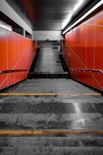 A staircase leading down with gray, wet steps and orange walls on both sides. The staircase has yellow safety lines near the edges and handrails attached to the walls. Fluorescent lighting runs along the top of the wall on the right side.