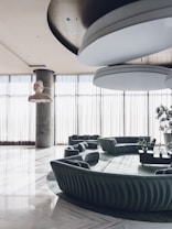 The interior features a contemporary, spacious seating area with a circular sofa and several cushions, positioned on a large rug. Natural light floods the room through floor-to-ceiling windows, partially covered by sheer curtains. A unique, artistic sculpture of a head is suspended from the ceiling. The overall design is modern and sophisticated.