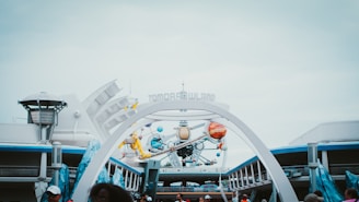 A futuristic theme park entrance featuring signage reading 'Tomorrowland' amidst art installations resembling planets and space-themed elements. The architecture suggests a vision of the future, with sleek lines and metallic accents. The scene is bustling with people, indicating a lively and active environment.