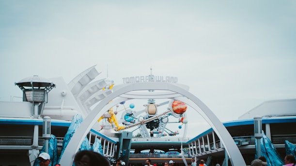 A futuristic theme park entrance featuring signage reading 'Tomorrowland' amidst art installations resembling planets and space-themed elements. The architecture suggests a vision of the future, with sleek lines and metallic accents. The scene is bustling with people, indicating a lively and active environment.