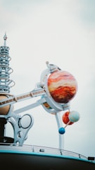 A futuristic structure with a model of a solar system, featuring large, colorful planet spheres mounted on metallic arms. The planets are intricately detailed, and the structure includes a series of mechanical and spiral elements, creating a sense of modernity and innovation against a cloudy sky.