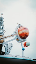 A futuristic structure with a model of a solar system, featuring large, colorful planet spheres mounted on metallic arms. The planets are intricately detailed, and the structure includes a series of mechanical and spiral elements, creating a sense of modernity and innovation against a cloudy sky.