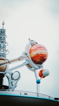 A futuristic structure with a model of a solar system, featuring large, colorful planet spheres mounted on metallic arms. The planets are intricately detailed, and the structure includes a series of mechanical and spiral elements, creating a sense of modernity and innovation against a cloudy sky.