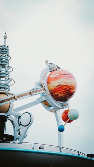 A futuristic structure with a model of a solar system, featuring large, colorful planet spheres mounted on metallic arms. The planets are intricately detailed, and the structure includes a series of mechanical and spiral elements, creating a sense of modernity and innovation against a cloudy sky.