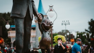A bronze statue of a man holding hands with a cartoon mouse character, located in an outdoor theme park setting with a castle-like structure in the background. People are seen walking and taking photographs, contributing to a lively and playful atmosphere.