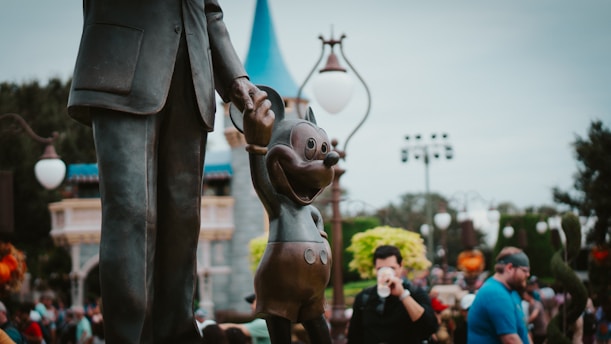 A bronze statue of a man holding hands with a cartoon mouse character, located in an outdoor theme park setting with a castle-like structure in the background. People are seen walking and taking photographs, contributing to a lively and playful atmosphere.