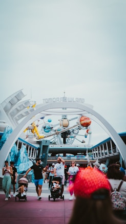 An amusement park entrance features futuristic architecture and a sign reading 'Tomorrowland'. People are walking beneath the structure, including a man pushing a stroller and others leisurely strolling around. In the background, a ride with colorful planet-like structures is visible, contributing to the theme park's sci-fi ambiance.