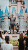 A bronze statue features a man in a suit holding hands with a cartoon mouse, set in front of a fairy-tale castle with blue turrets. A crowd of visitors, some wearing Mickey Mouse ears, gather around the statue, adding a lively atmosphere.