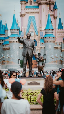 A bronze statue features a man in a suit holding hands with a cartoon mouse, set in front of a fairy-tale castle with blue turrets. A crowd of visitors, some wearing Mickey Mouse ears, gather around the statue, adding a lively atmosphere.