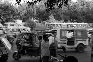 A busy street scene featuring multiple auto rickshaws and a bus in motion, with trees and buildings in the background. A man stands in the foreground using a cellphone.