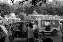 A busy street scene featuring multiple auto rickshaws and a bus in motion, with trees and buildings in the background. A man stands in the foreground using a cellphone.
