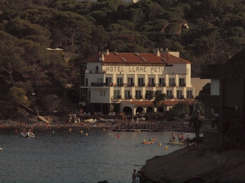 A multi-story coastal hotel named 'Hotel Llane Petit' with a red-tiled roof, situated by a water edge. Several people are seen on the shore and in the water engaged in various activities. The background is filled with dense greenery and trees.