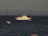 A large white yacht with multiple decks is anchored in the ocean, surrounded by several smaller sailboats. Yellow buoys form a line in the foreground, creating a path toward the yacht. The sky is overcast, giving the scene a calm and serene atmosphere.