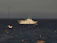 A large white yacht with multiple decks is anchored in the ocean, surrounded by several smaller sailboats. Yellow buoys form a line in the foreground, creating a path toward the yacht. The sky is overcast, giving the scene a calm and serene atmosphere.
