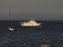 A large white yacht with multiple decks is anchored in the ocean, surrounded by several smaller sailboats. Yellow buoys form a line in the foreground, creating a path toward the yacht. The sky is overcast, giving the scene a calm and serene atmosphere.