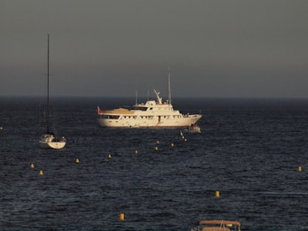 A large white yacht with multiple decks is anchored in the ocean, surrounded by several smaller sailboats. Yellow buoys form a line in the foreground, creating a path toward the yacht. The sky is overcast, giving the scene a calm and serene atmosphere.