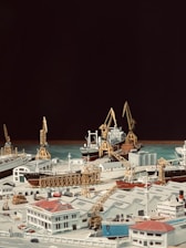 A detailed model of a bustling port scene, featuring various cargo ships docked at piers with towering cranes poised for loading and unloading. The foreground includes model buildings with distinctive architecture, matching the maritime theme with an array of smaller details such as trucks and stacked containers. The background contrasts the detailed foreground with a solid dark backdrop, highlighting the model elements.