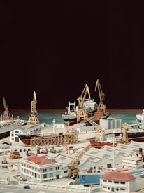 A detailed model of a bustling port scene, featuring various cargo ships docked at piers with towering cranes poised for loading and unloading. The foreground includes model buildings with distinctive architecture, matching the maritime theme with an array of smaller details such as trucks and stacked containers. The background contrasts the detailed foreground with a solid dark backdrop, highlighting the model elements.