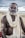 An older man with a beard and traditional markings on his forehead and arms wears a white garment and sits against a backdrop of a cloudy sky and sandy beach.