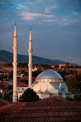 A mosque with two tall white minarets and a large dome is situated among a cluster of homes with red-tiled roofs. The setting appears to be a small town or village, surrounded by rolling hills, under a cloudy sky during sunset or sunrise.