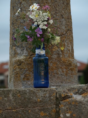 A vibrant blue glass bottle filled with an assortment of colorful wildflowers including daisies and other blooms. The bottle is perched on a weathered stone surface with the backdrop of a stone pillar. The image conveys an outdoor setting with partially visible buildings in the background.