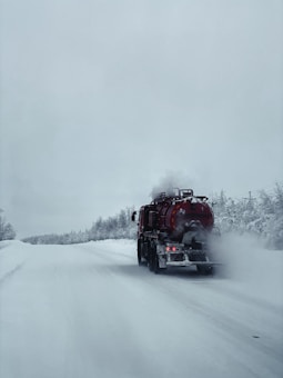 A tanker truck is driving on a snow-covered road surrounded by trees. The sky is overcast, creating a cold and isolated atmosphere. The truck's back lights are on, and there is visible steam or exhaust coming from it, blending with the snowy environment.