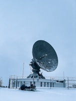 A large satellite dish is positioned in a snow-covered landscape, enclosed by a metal fence. The sky is overcast, contributing to the cold and isolated atmosphere. In the foreground, there is a person on a snowmobile, dressed in winter gear, likely indicating a remote or cold location.