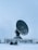 A large satellite dish is positioned in a snow-covered landscape, enclosed by a metal fence. The sky is overcast, contributing to the cold and isolated atmosphere. In the foreground, there is a person on a snowmobile, dressed in winter gear, likely indicating a remote or cold location.