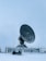 A large satellite dish is positioned in a snow-covered landscape, enclosed by a metal fence. The sky is overcast, contributing to the cold and isolated atmosphere. In the foreground, there is a person on a snowmobile, dressed in winter gear, likely indicating a remote or cold location.