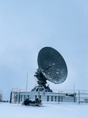 A large satellite dish is positioned in a snow-covered landscape, enclosed by a metal fence. The sky is overcast, contributing to the cold and isolated atmosphere. In the foreground, there is a person on a snowmobile, dressed in winter gear, likely indicating a remote or cold location.