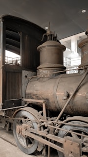 A vintage steam locomotive featuring large metal wheels, a cylindrical boiler, and distinct mechanical components. The metal surfaces appear aged with rust and wear, suggesting historical significance. The background includes industrial elements such as metal beams and fluorescent lighting.