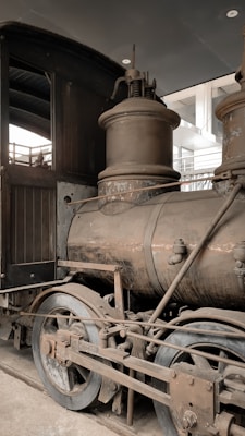 A vintage steam locomotive with a weathered appearance, showing detailed components like the boiler, wheels, and connecting rods. The setting suggests an indoor environment, possibly a museum or historical display.