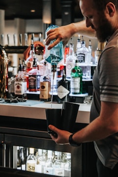 A bartender is pouring a drink using a blue-tinted bottle into two black cocktail shakers. The background is filled with various bottles of liquor neatly arranged on a bar counter.