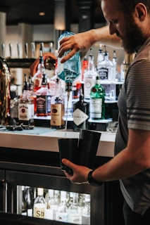 A bartender is pouring a drink using a blue-tinted bottle into two black cocktail shakers. The background is filled with various bottles of liquor neatly arranged on a bar counter.