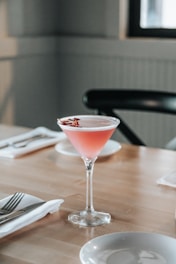 A pink cocktail garnished with flower petals is served in a martini glass on a wooden table. The table is set with a folded white napkin and cutlery, and the background includes a black chair and subtle natural lighting.