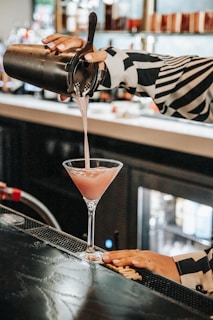 A person is pouring a pink cocktail from a metal shaker into a martini glass at a dimly lit bar counter. The background is slightly out of focus, showing shelves lined with bottles, creating a cozy and stylish atmosphere.
