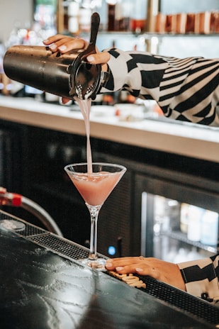 A person is pouring a pink cocktail from a metal shaker into a martini glass at a dimly lit bar counter. The background is slightly out of focus, showing shelves lined with bottles, creating a cozy and stylish atmosphere.