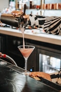 A person is pouring a pink cocktail from a shaker into a martini glass on a bar counter. The bartender wears a black and white patterned shirt, and the bar area is dimly lit with bottles in the background.