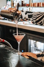 A person is pouring a pink cocktail from a shaker into a martini glass on a bar counter. The bartender wears a black and white patterned shirt, and the bar area is dimly lit with bottles in the background.