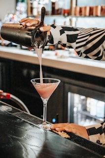 A person is pouring a pink cocktail from a shaker into a martini glass on a bar counter. The bartender wears a black and white patterned shirt, and the bar area is dimly lit with bottles in the background.