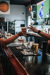 In a bar setting, two people are depicted exchanging a drink across a counter. Various beverages in different types of glasses are arranged on the counter, including cocktails and beer. The background features a blurry TV screen displaying a sports event with a player in blue uniform. The environment is cozy and dimly lit, and there are candles and bottles in the background.