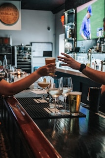 In a bar setting, two people are depicted exchanging a drink across a counter. Various beverages in different types of glasses are arranged on the counter, including cocktails and beer. The background features a blurry TV screen displaying a sports event with a player in blue uniform. The environment is cozy and dimly lit, and there are candles and bottles in the background.