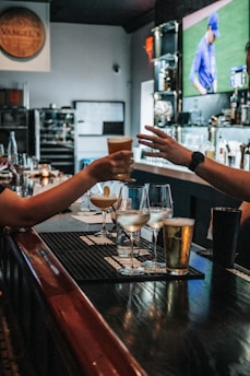 In a bar setting, two people are depicted exchanging a drink across a counter. Various beverages in different types of glasses are arranged on the counter, including cocktails and beer. The background features a blurry TV screen displaying a sports event with a player in blue uniform. The environment is cozy and dimly lit, and there are candles and bottles in the background.