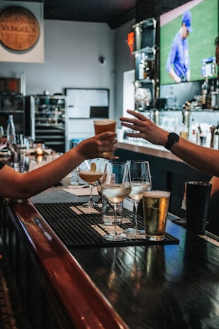 In a bar setting, two people are depicted exchanging a drink across a counter. Various beverages in different types of glasses are arranged on the counter, including cocktails and beer. The background features a blurry TV screen displaying a sports event with a player in blue uniform. The environment is cozy and dimly lit, and there are candles and bottles in the background.
