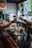 In a bar setting, two people are depicted exchanging a drink across a counter. Various beverages in different types of glasses are arranged on the counter, including cocktails and beer. The background features a blurry TV screen displaying a sports event with a player in blue uniform. The environment is cozy and dimly lit, and there are candles and bottles in the background.